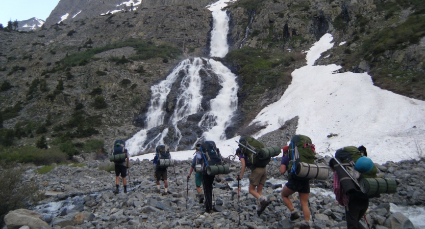 A group of people carrying backpacks hike across rocky terrain near a frozen waterfall. 